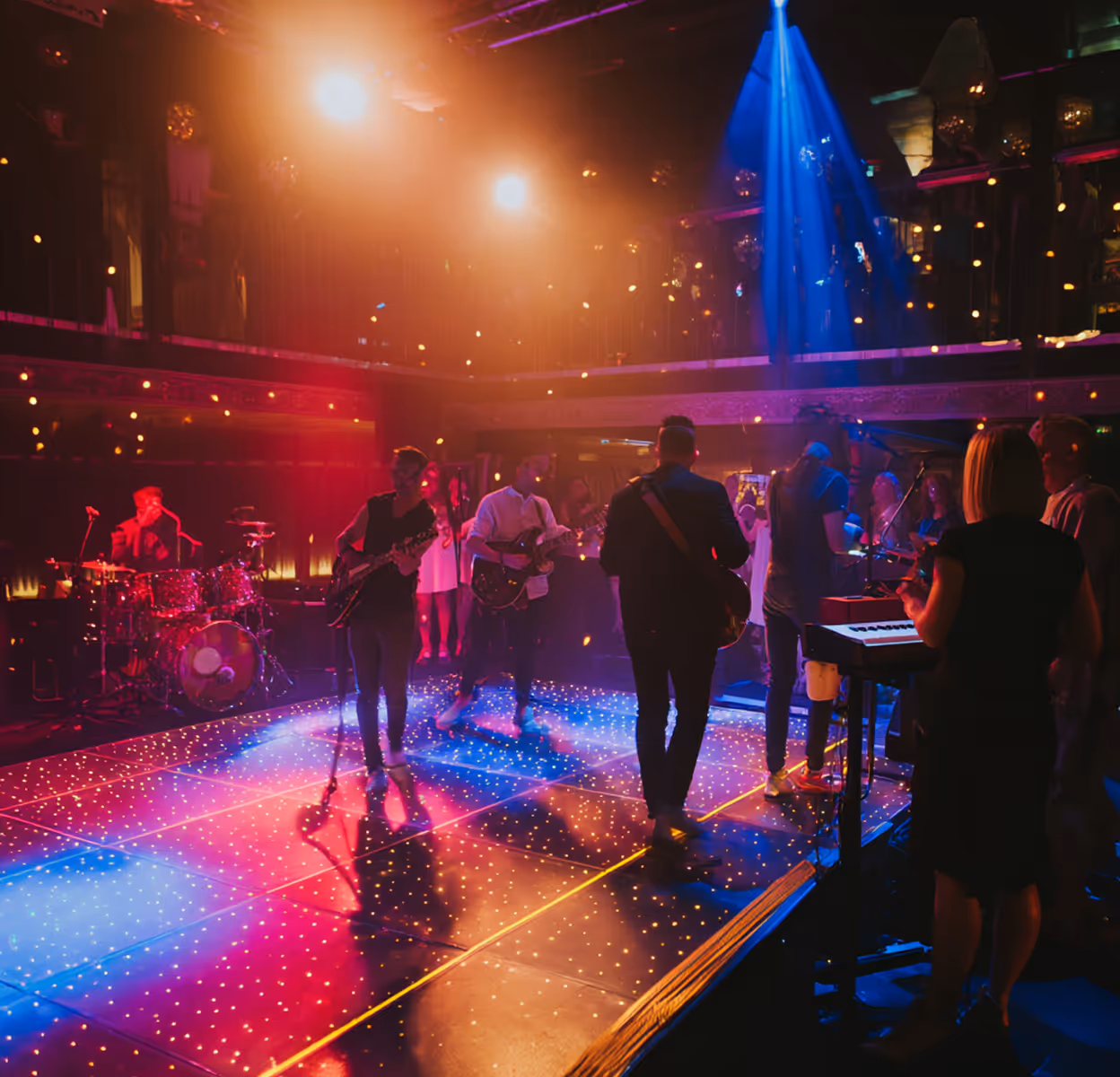 Band performing on a stage with colorful lights and a drummer, guitarists, and keyboard player in a lively concert venue.