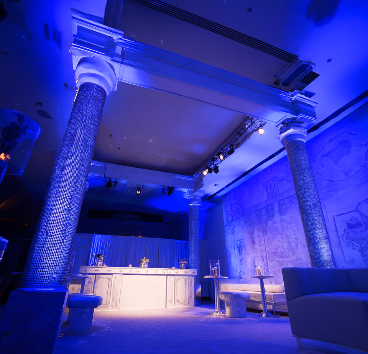 Interior of a dimly lit modern lounge with blue lighting, white columns, a bar counter, and seating areas.