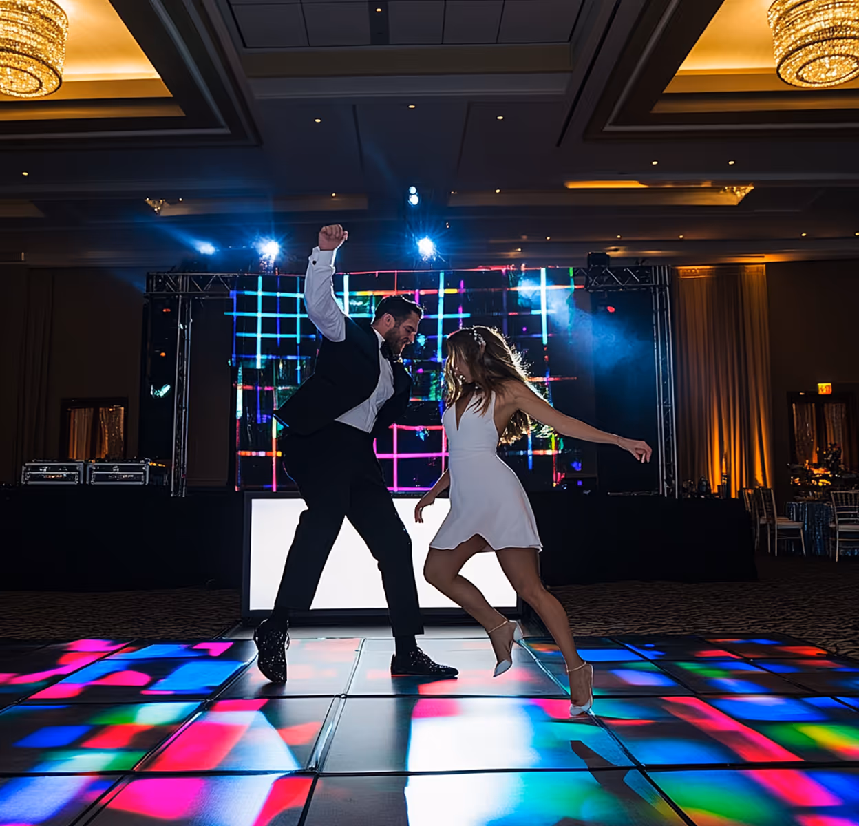 Man in black suit and woman in white dress dancing energetically on a colorful illuminated dance floor at an indoor event.