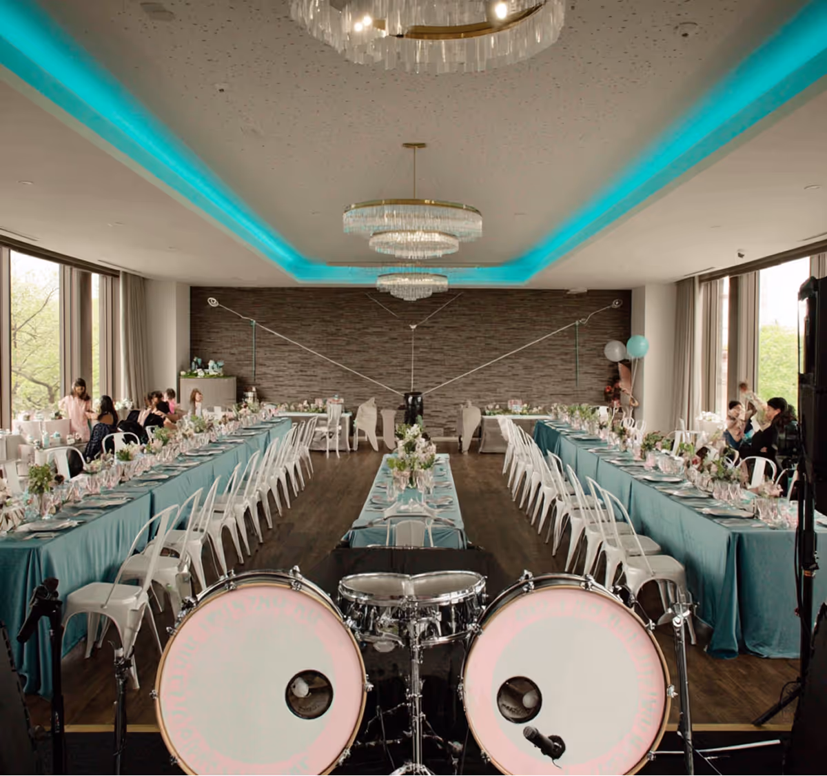 A long dining room set for an event with teal tablecloths, white chairs, floral centerpieces, and a drum set in the foreground.