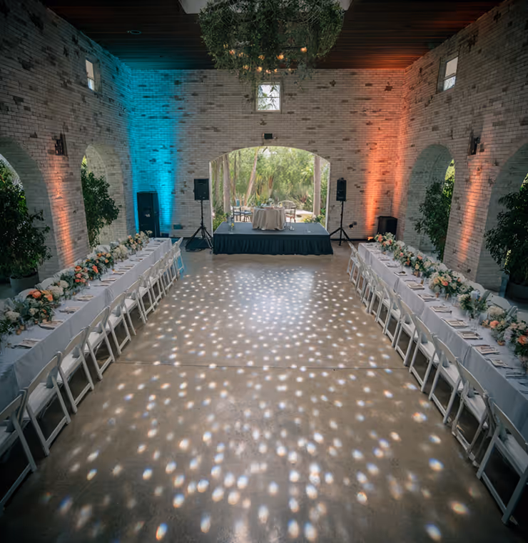 Elegant event space with two long banquet tables set with white chairs, floral centerpieces, and soft blue and orange uplighting, facing a small stage under a hanging greenery chandelier.