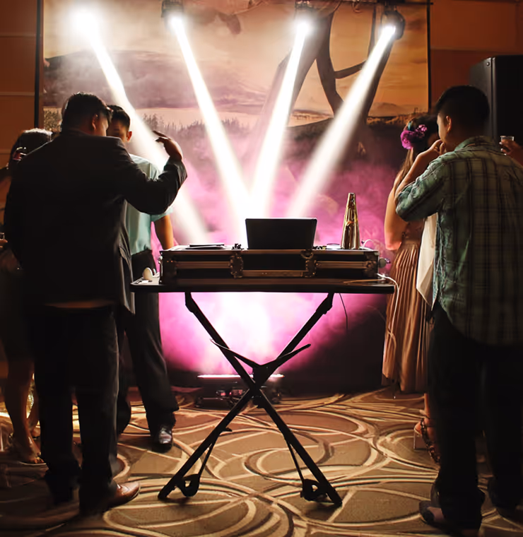 People standing around a DJ booth with bright white stage lights and pink fog in the background at an indoor event.