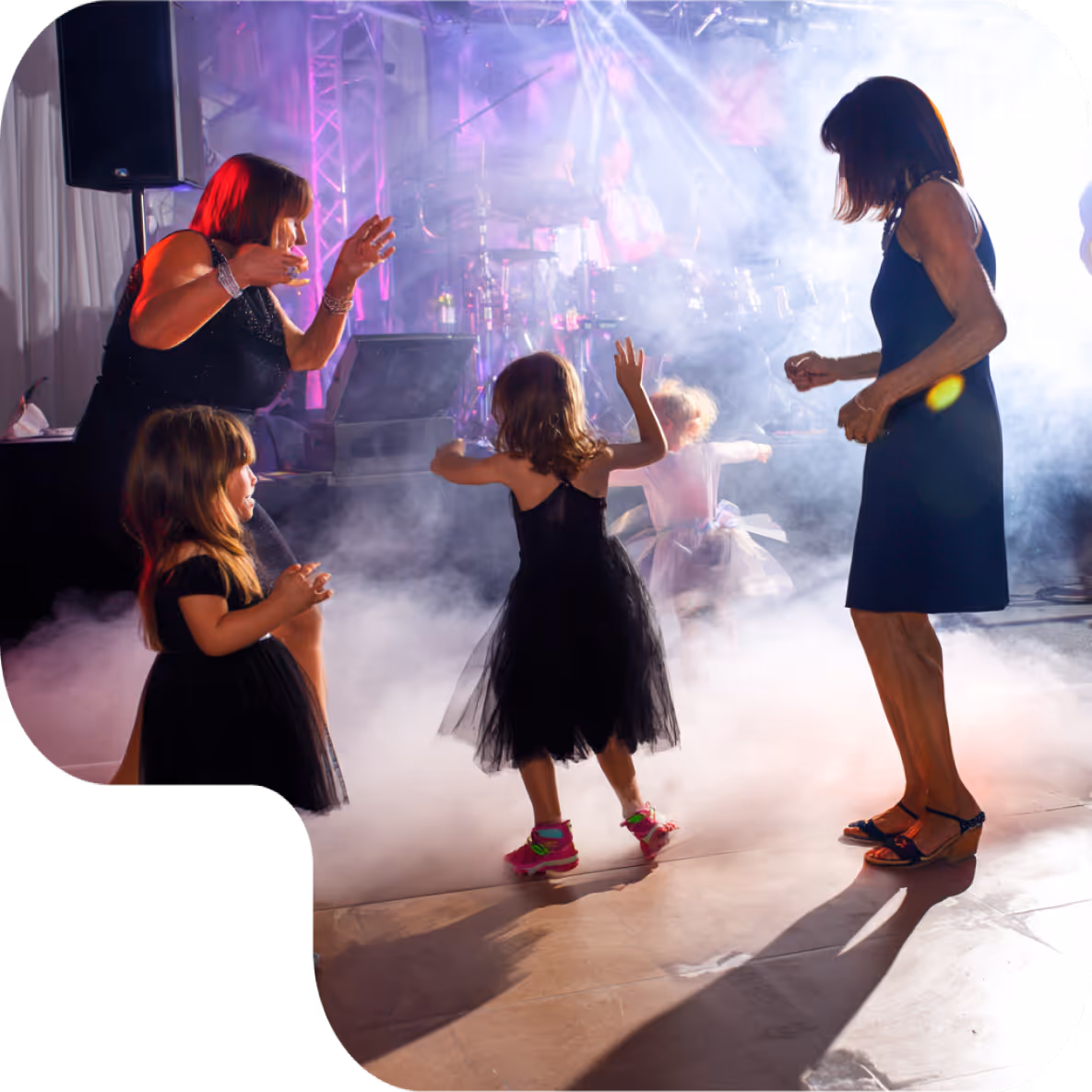 Two women and three young girls dancing on a foggy dance floor with stage lights and a drummer in the background.