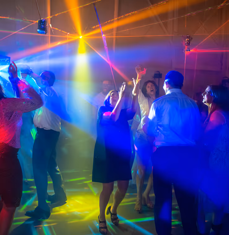 Group of people dancing and raising hands amid colorful stage lights and laser effects at an indoor party.