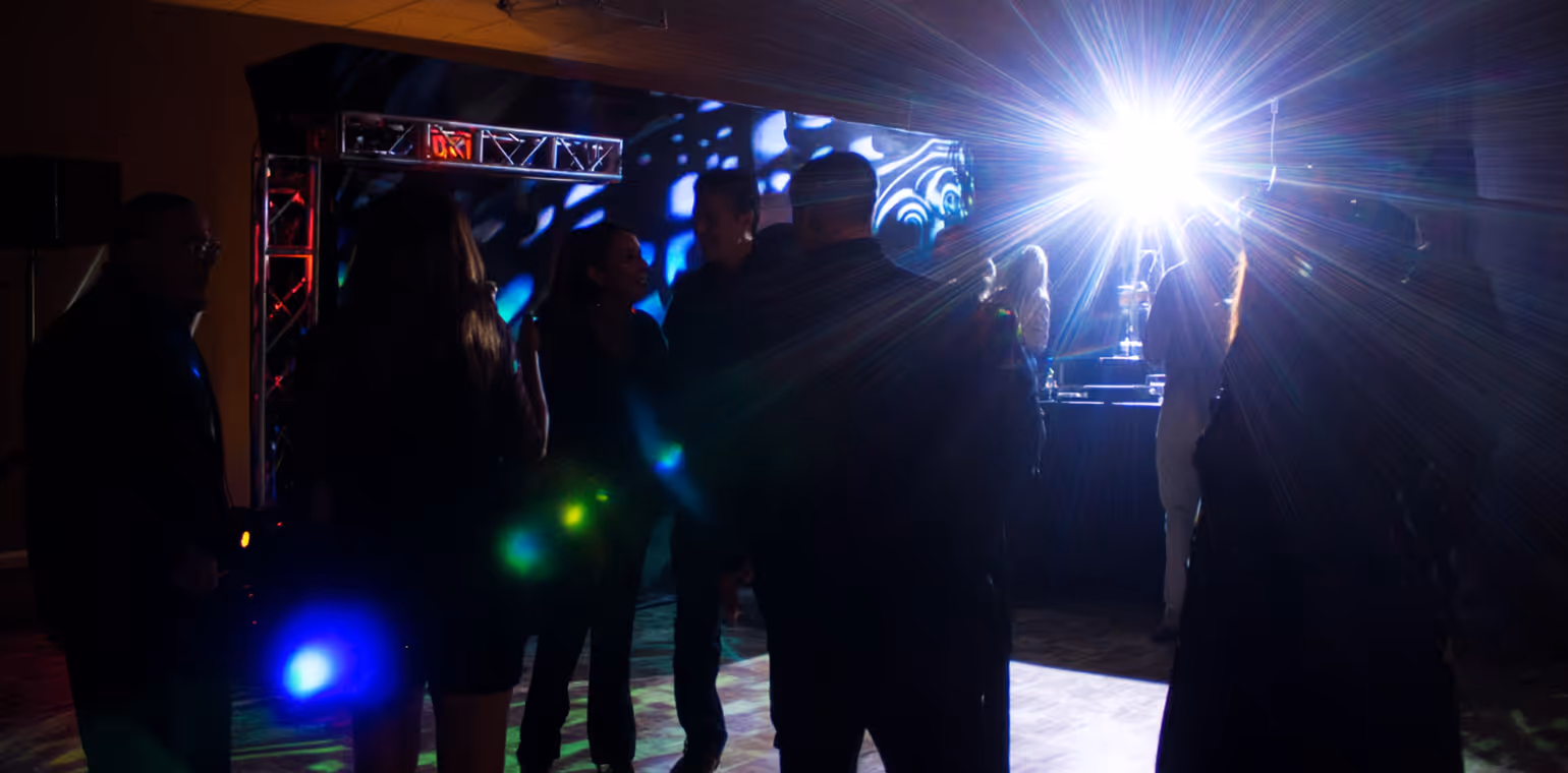Silhouettes of people dancing in a dark room with bright stage lights and colorful spotlights in the background.