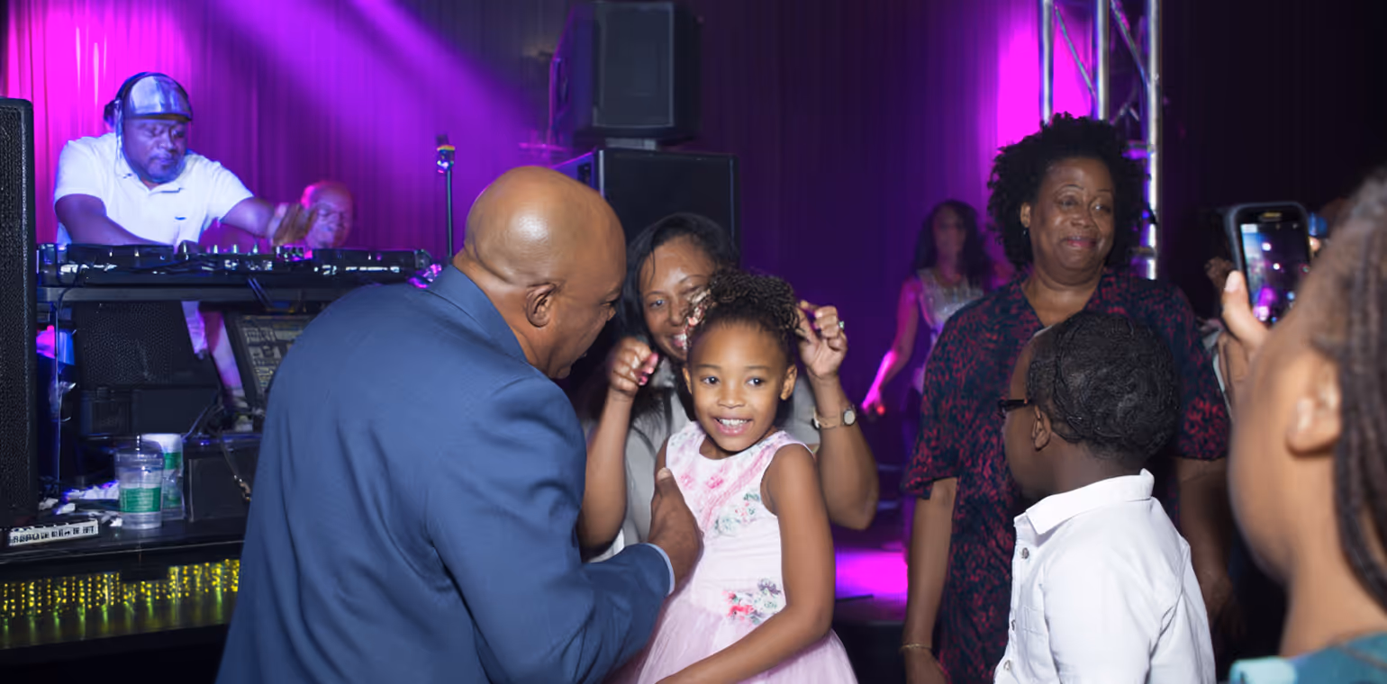 A smiling young girl in a pink dress dancing with a man in a blue suit and surrounded by adults at a party with a DJ in the background under purple lighting.