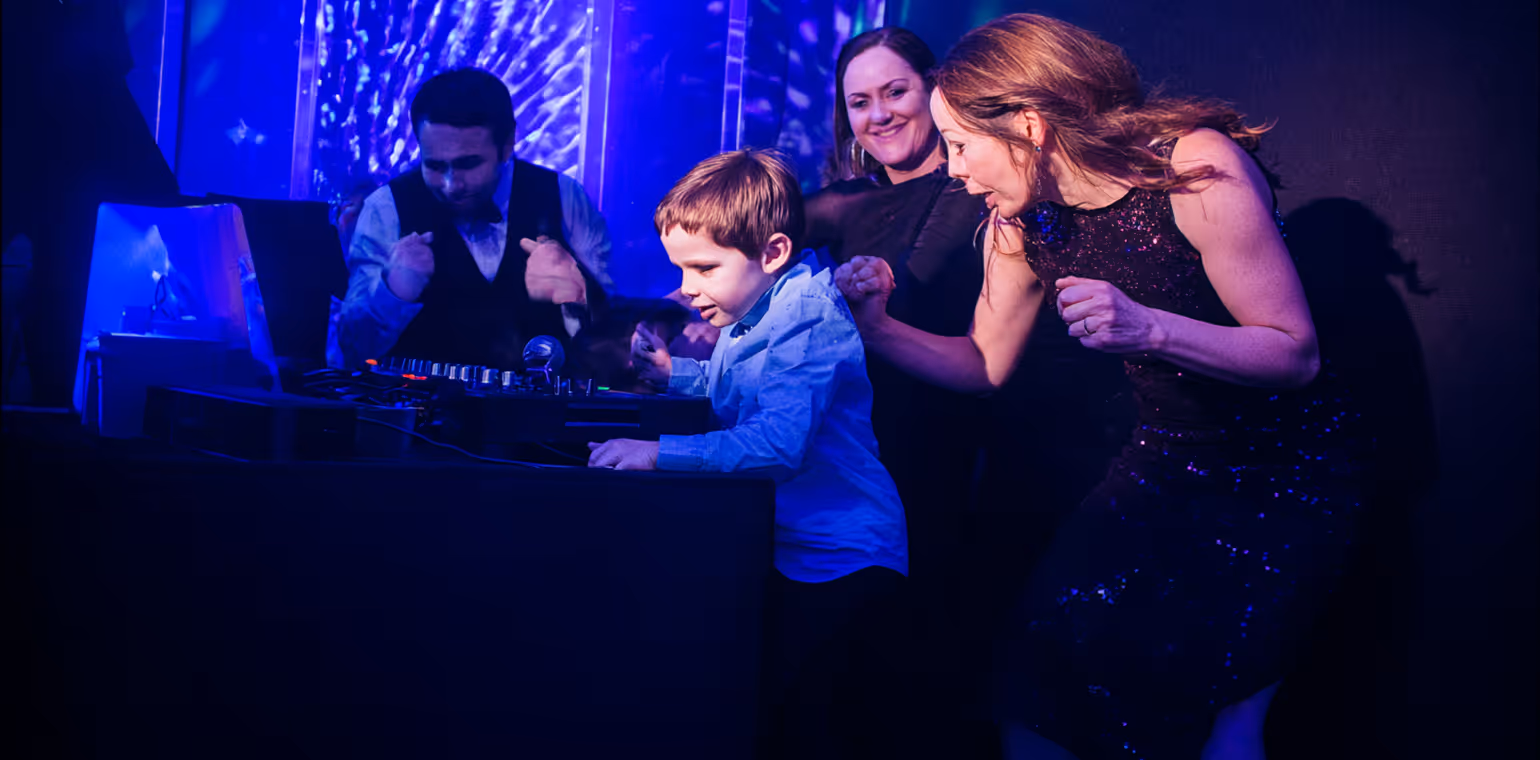 A young boy operates DJ equipment while two women and a man enjoy and cheer near him in a dark, blue-lit party setting.