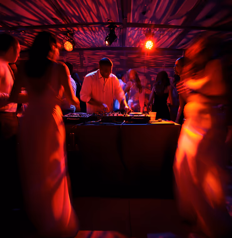DJ wearing a white shirt mixing music at a nightclub with people dancing around under red and purple lighting.