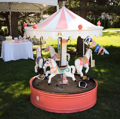 Small vintage carousel with four white horses on a red base set on green grass in a garden area.
