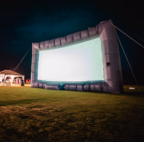 Large inflatable outdoor movie screen illuminated at night on a grassy field with a tent and people in the background.