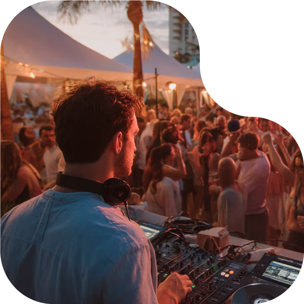 DJ wearing headphones plays music at an outdoor party with a crowd dancing under tents at dusk.