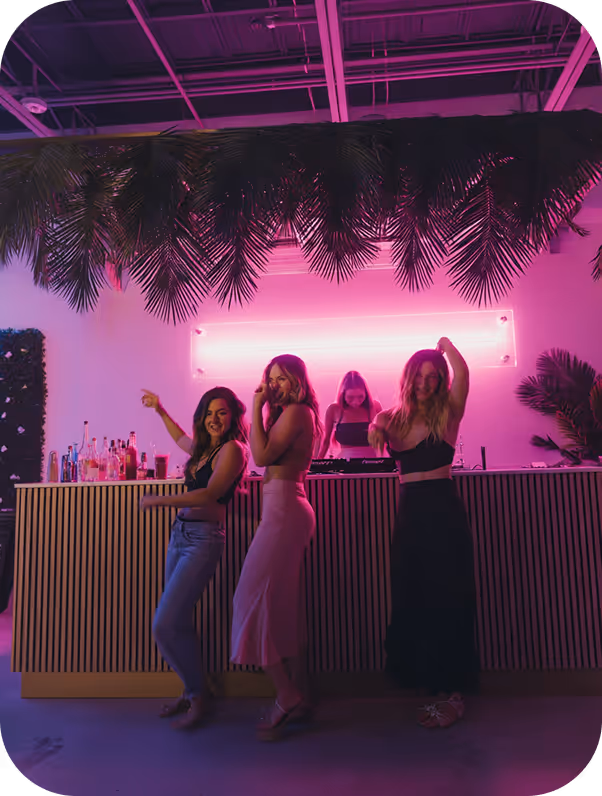 Three women dancing and posing in front of a bar with a DJ playing music under pink neon lighting and palm leaves overhead.