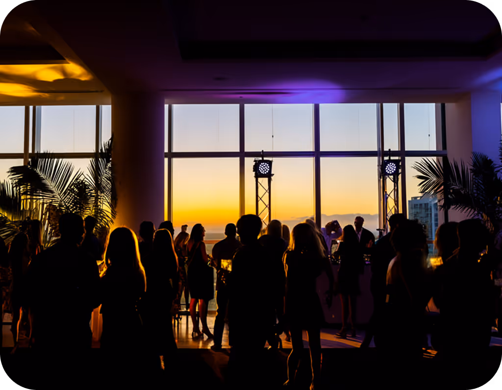 Silhouettes of people socializing indoors against large windows showing a sunset sky.