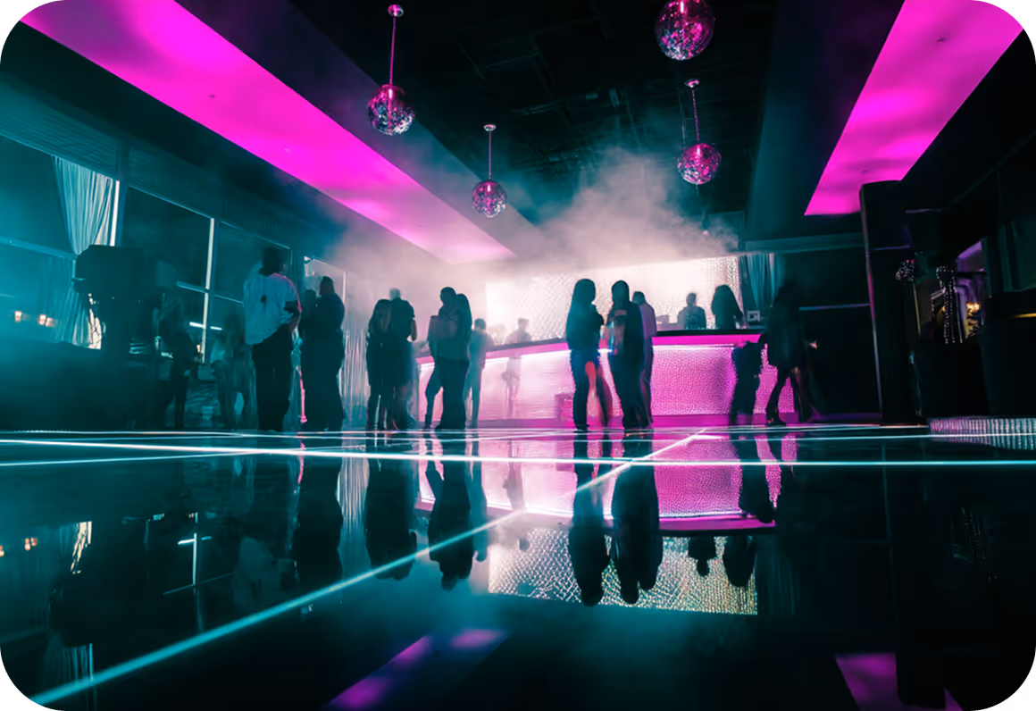 People socializing inside a club with neon pink and blue lighting and a reflective dance floor.