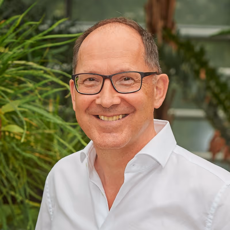 Ein Mann mit Brille und weißem Hemd vor einer grünen Hecke lächelt freundlich in die Kamera