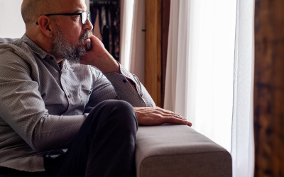 Older man with glasses and a beard sitting on a sofa by a window, looking outside thoughtfully.