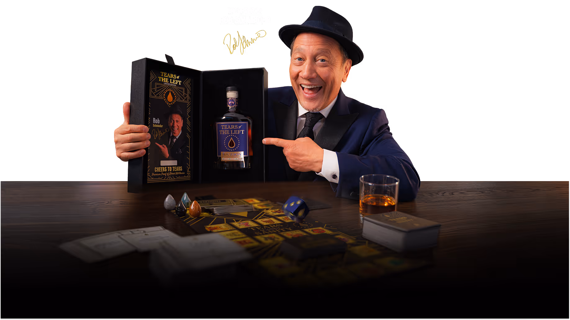 Rob Schneider in a suit and hat smiling while pointing to a box containing a bottle labeled 'Tears of the Left' bourbon on a table with board game pieces and a glass of whiskey.