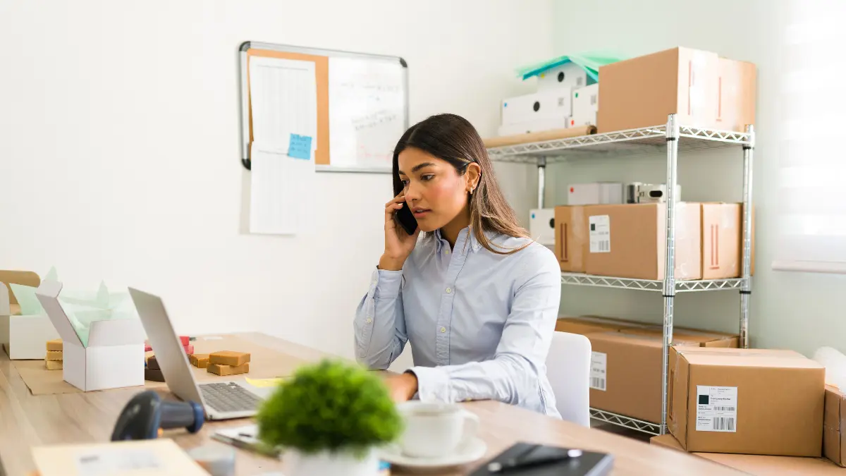 Office worker talking on the phone at her desk with shelves of boxes and supplies in the background