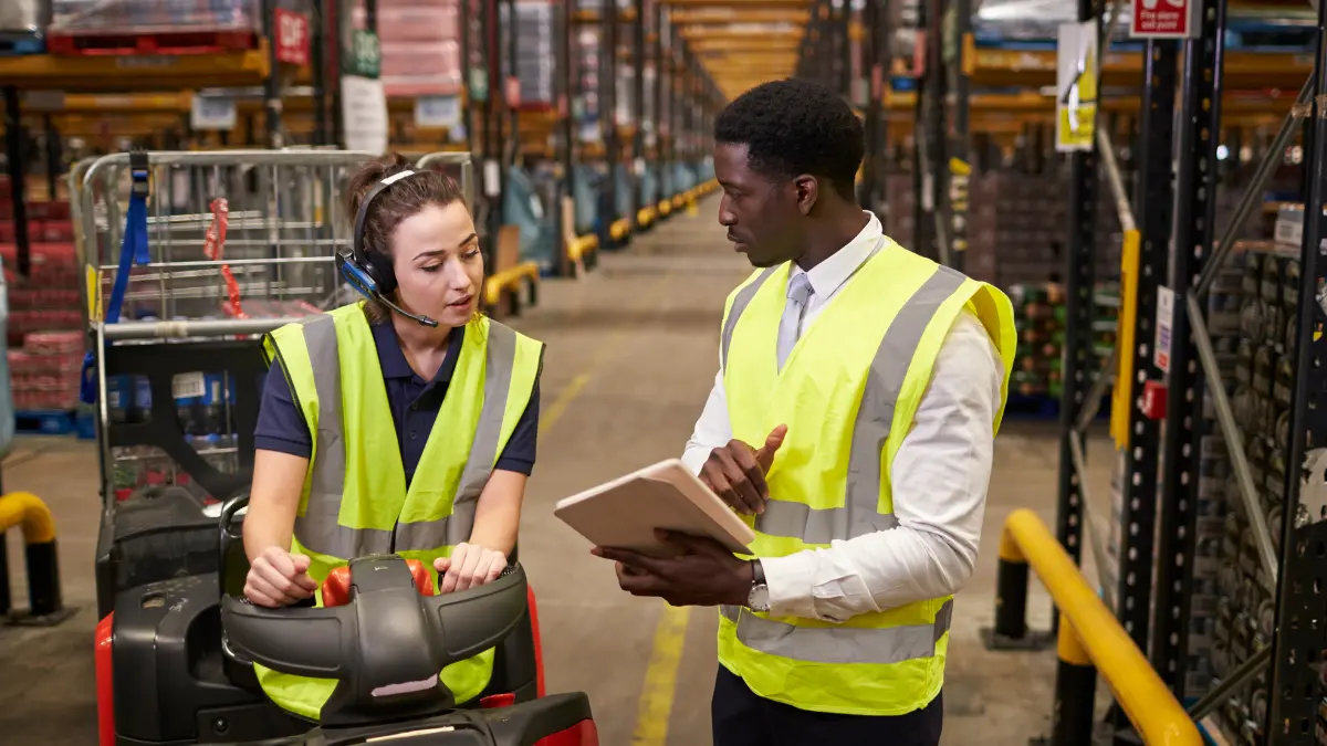 Supervisor in a safety vest holding a tablet, discussing with a forklift operator in a large warehouse