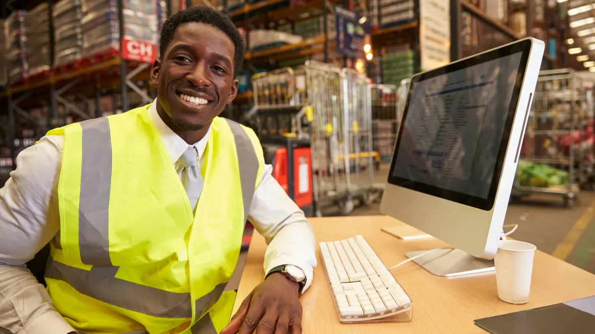 Warehouse manager in a safety vest smiling at his desk with a computer in a busy storage facility