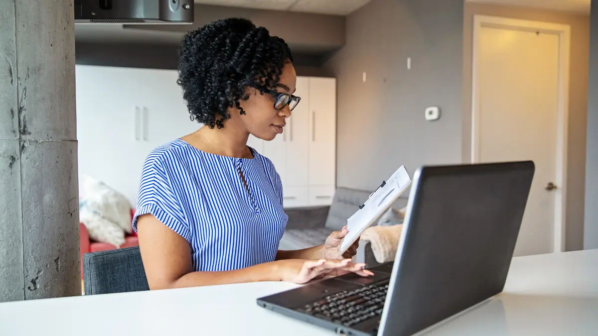 Automated risk management system being reviewed by a woman referencing a clipboard while working on a laptop at home