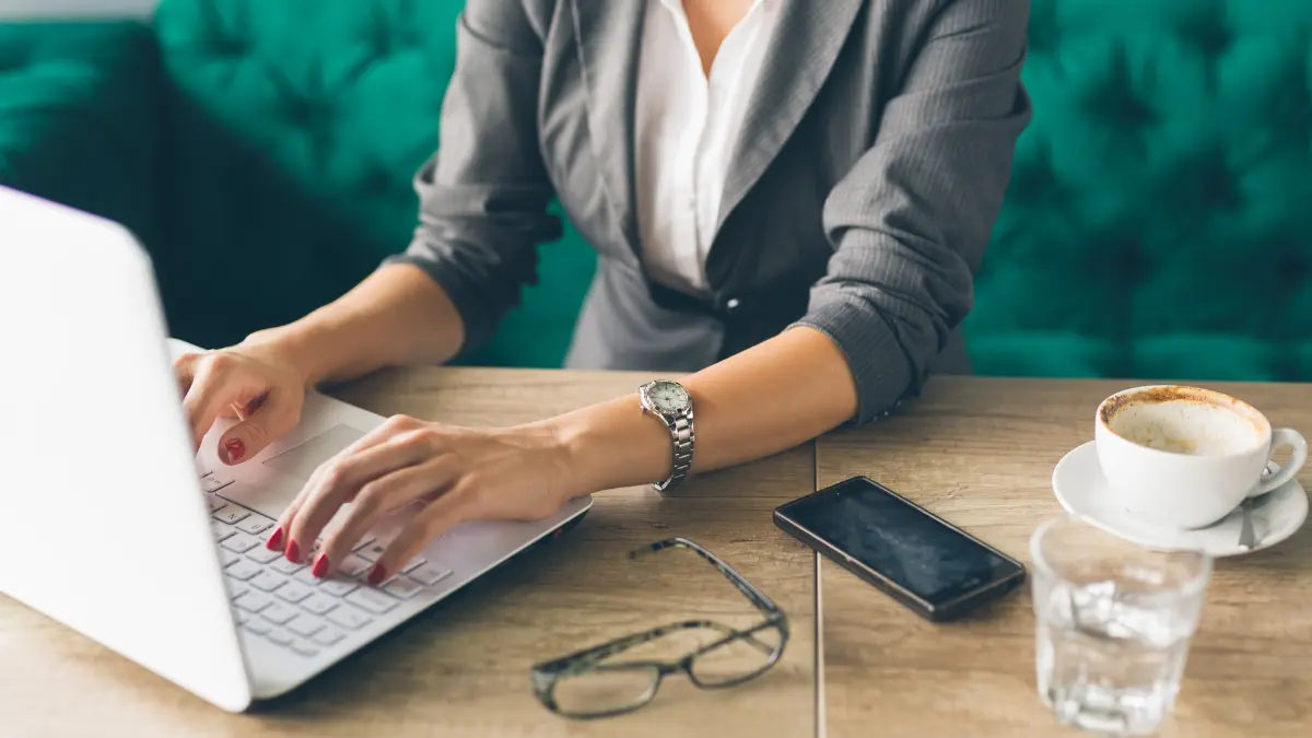 Risk management consultant in a blazer working on a laptop at a café table with coffee, phone, and glasses nearby.