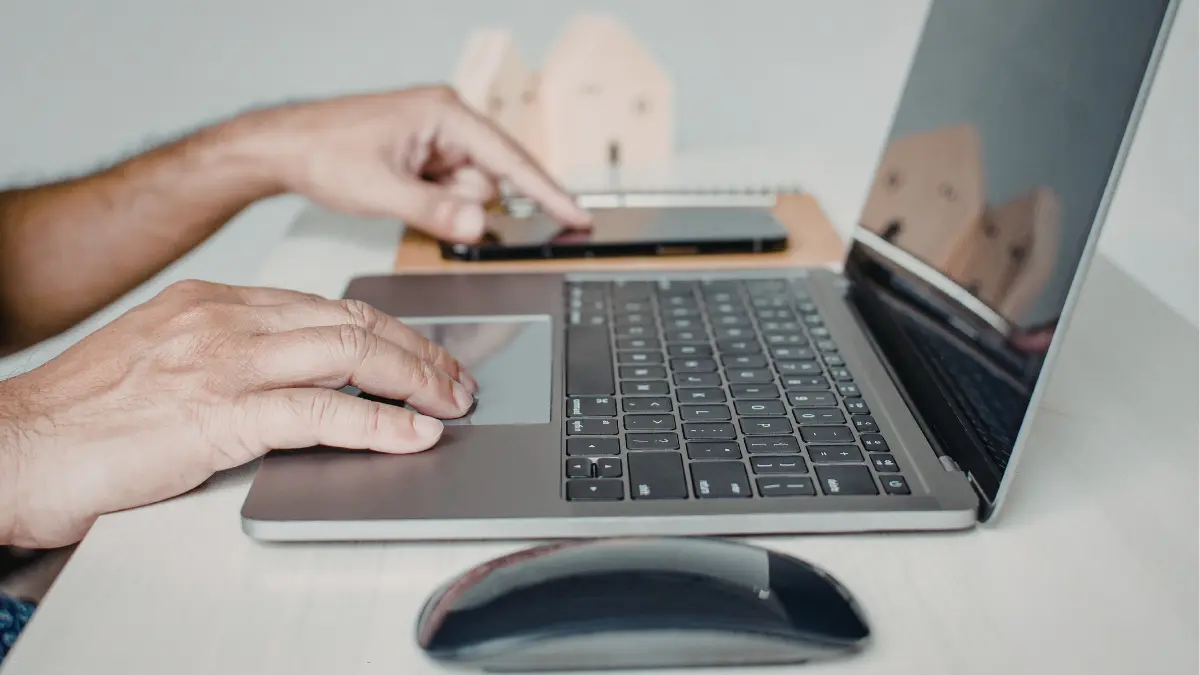 Enterprise risk assessment underway as a professional navigates a MacBook and smartphone side by side on a desk with a wireless mouse.