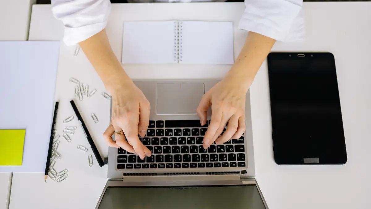 Risk management workflow captured from above showing hands typing on a laptop surrounded by a tablet, notebook, and scattered paper clips.
