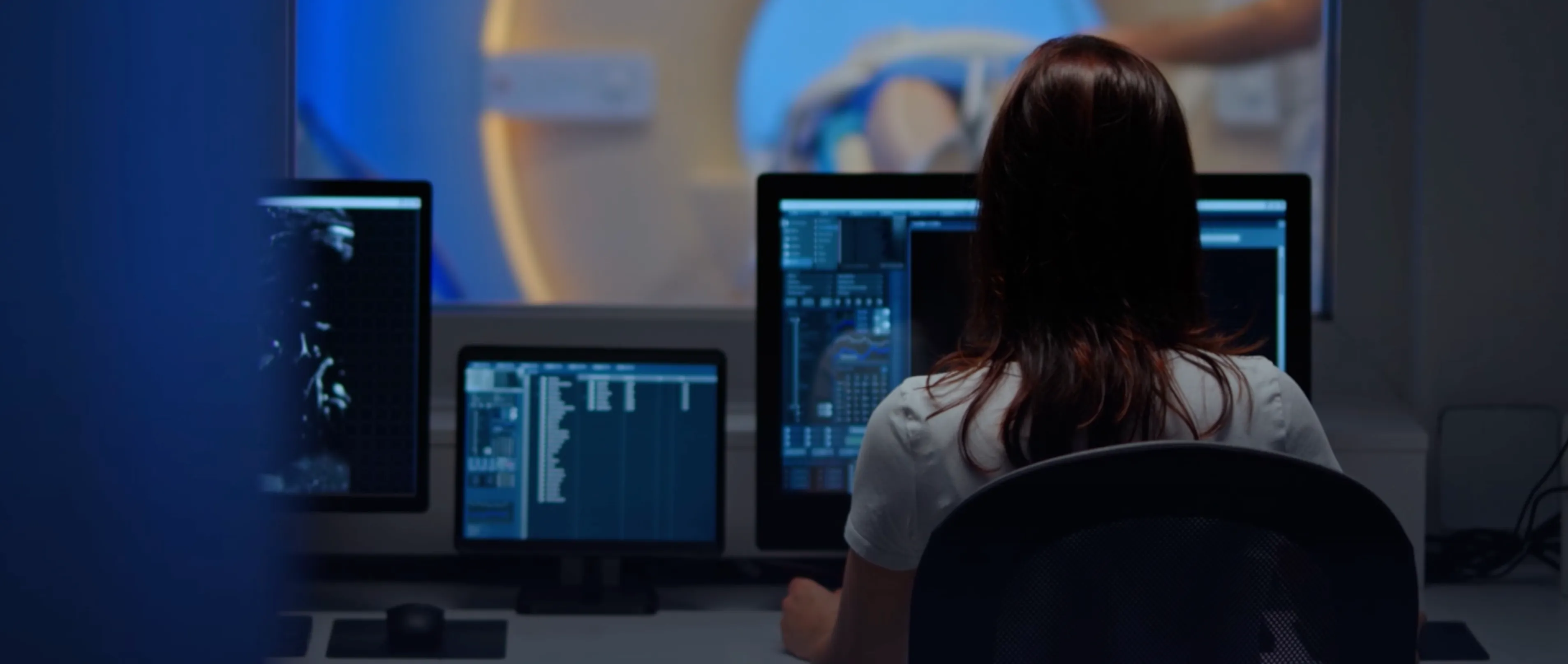A woman seated at a desk, focused on three computer screens displaying various data and information.