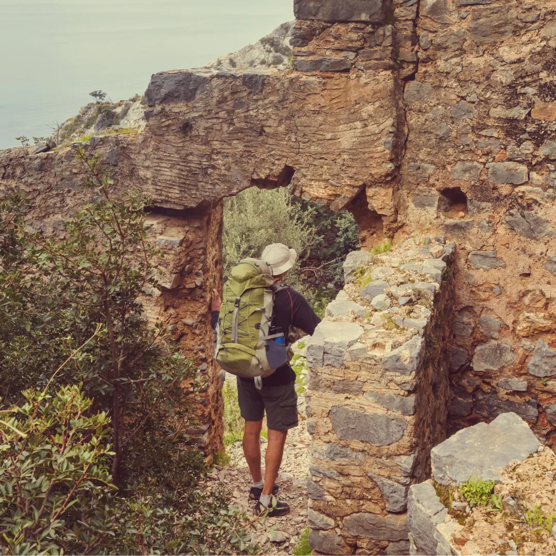 Hiker Exploring Ancient Ruins on the Lycian Way Trail A lone hiker with a green backpack walks through weathered stone ruins along the Lycian Way, overlooking the Mediterranean coast and forest.
