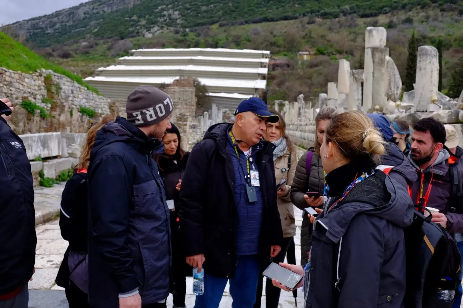 Tour Guide Leading Group in Ancient City of Ephesus A local guide explains historical ruins to a tour group in the ancient city of Ephesus, with stone columns and green hills in the background.