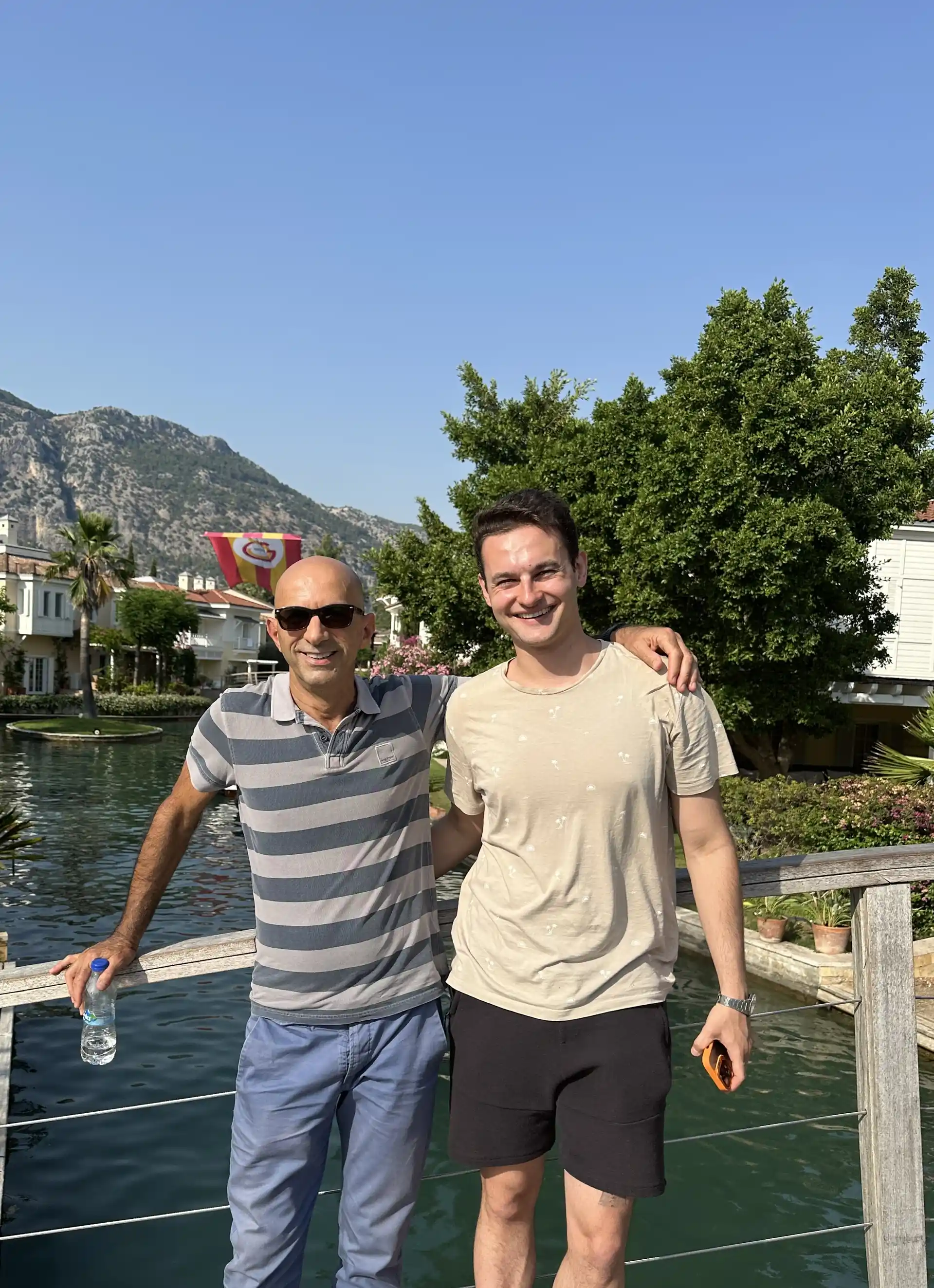 Two men standing by a waterfront marina with mountain backdrop in southwest Turkey, featuring calm water channels and Mediterranean coastal scenery.