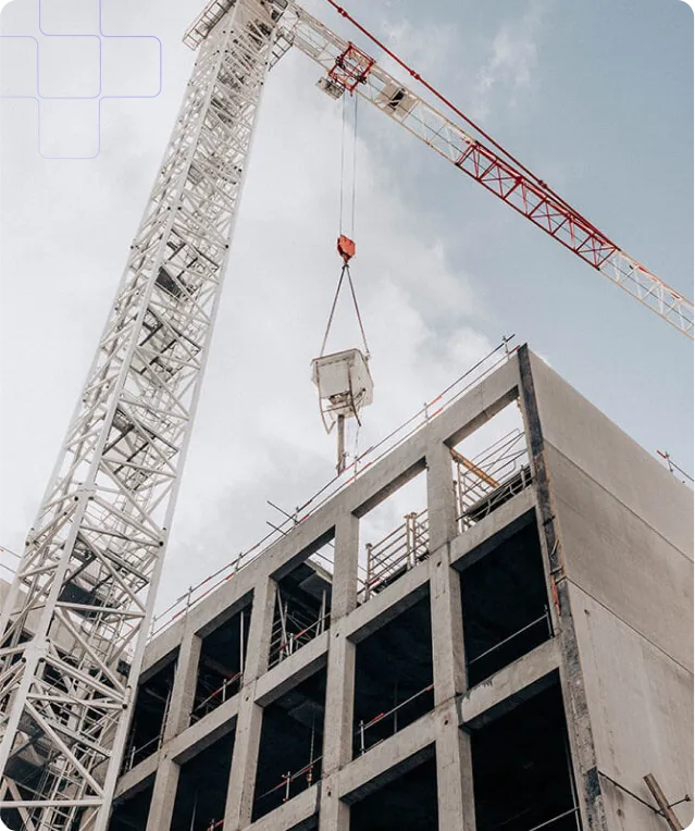 A low-angle shot shows a construction site with a tall crane lifting a container above a concrete building frame. The crane, predominantly white with red accents on its upper arm, extends high into the cloudy sky. The building frame, made of gray concrete, features a grid of empty window and door spaces, suggesting ongoing construction. Scaffolding and safety barriers are visible around the building's edges. The overall scene conveys a sense of progress and industry.