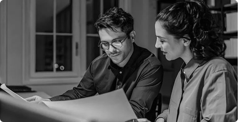A black and white image shows a man and a woman looking at a document. The man is on the left and is wearing glasses and a button-down shirt. The woman is on the right and has her hair pulled back. They are both sitting at a table, and there is a window and a bookshelf in the background.