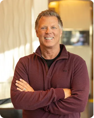 A portrait of a smiling middle-aged man with short gray hair, wearing a maroon long-sleeved shirt, standing with his arms crossed.
