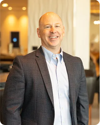 A smiling man with a bald head, wearing a blue button-down shirt and a gray plaid blazer, stands indoors.