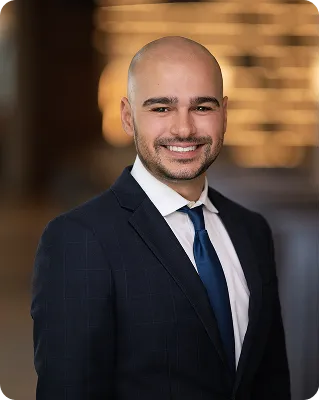 A professional headshot of a smiling man with a shaved head, wearing a navy blue suit, white shirt, and blue tie. The background is blurred.