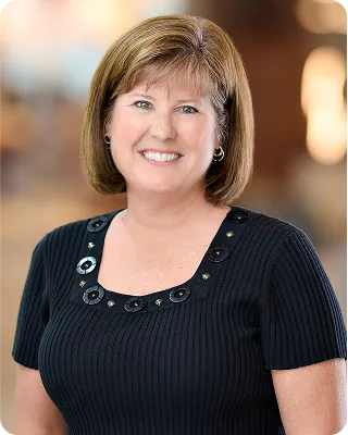 A headshot of a smiling woman with short brown hair, wearing a black top with decorative buttons around the neckline. She has fair skin and is wearing gold hoop earrings. The background is blurred, suggesting an indoor setting.