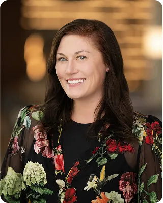 A professional headshot of a smiling woman with long brown hair wearing a black top and a sheer black floral print jacket. The background is blurred.