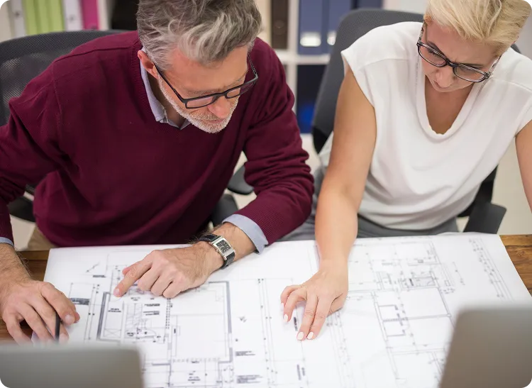 Two people are looking at a blueprint. The man on the left is wearing a maroon sweater and glasses, and he is pointing at the blueprint with his right index finger. The woman on the right is wearing a white shirt and glasses, and she has her right hand on the blueprint. There are two laptops in the foreground, and some binders in the background.