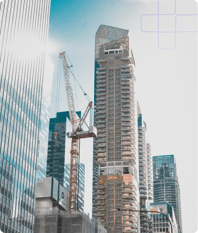 A low-angle shot of a city skyline with several skyscrapers and a construction crane. The buildings are modern and made of glass and steel. The crane is orange and white and is positioned in front of one of the buildings. The sky is blue with some clouds. There is a faint blue plus sign graphic in the upper right corner.