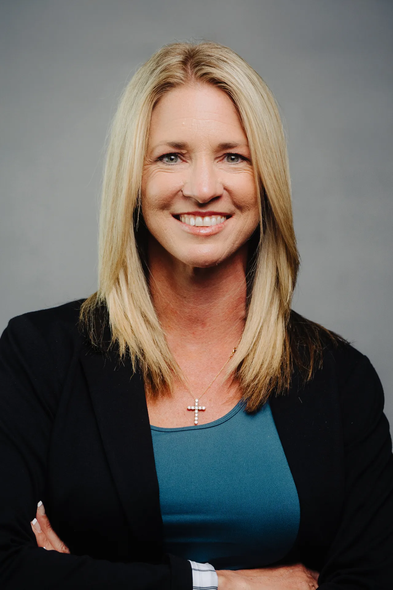 A professional headshot of a blonde woman with blue eyes smiling at the camera. She is wearing a black blazer over a teal top and a gold necklace with a cross pendant. Her arms are crossed. The background is a solid gray color.