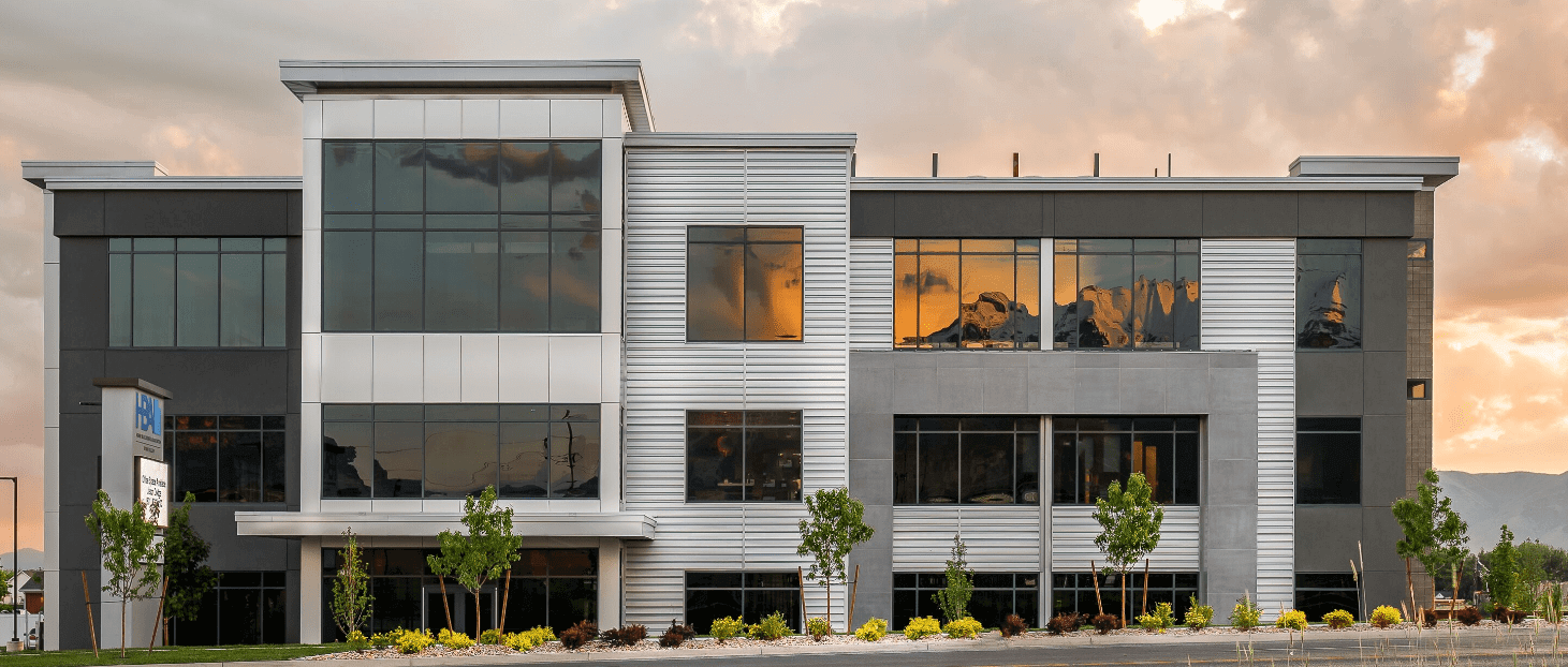 Modern commercial building with large reflective windows showing sunset sky, surrounded by small trees and landscaping.