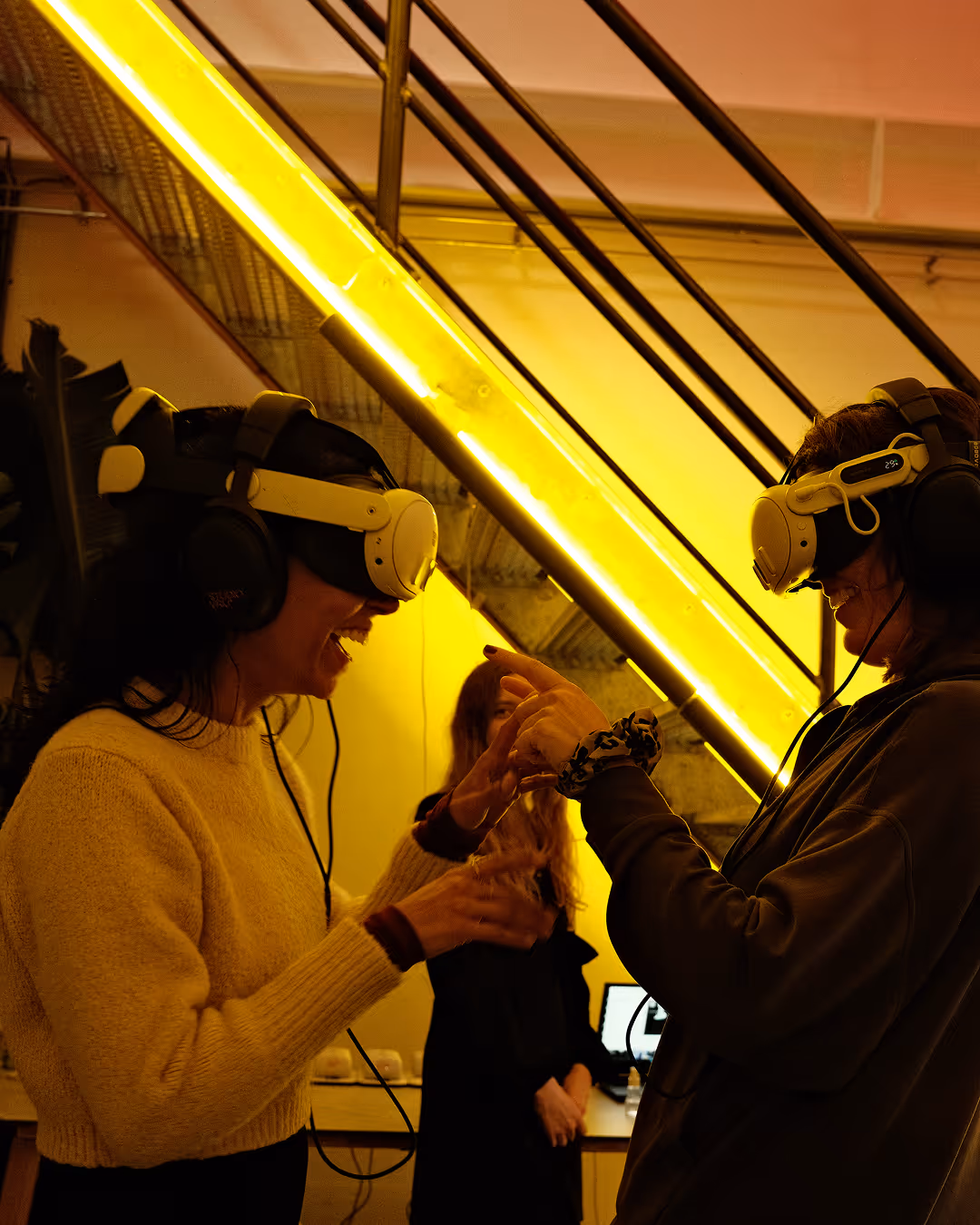 Two participants in VR headsets interacting with hand tracking under yellow neon lighting at ADE premiere of Show Me the Light VR Silent Disco.