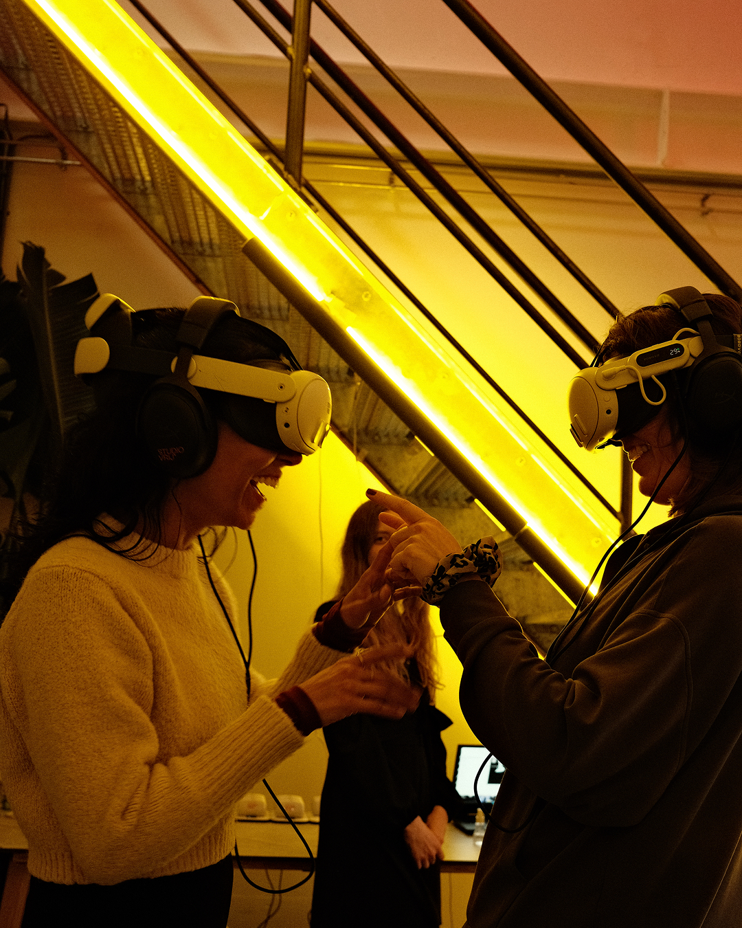Two participants in VR headsets interacting with hand tracking under yellow neon lighting at ADE premiere of Show Me the Light VR Silent Disco.