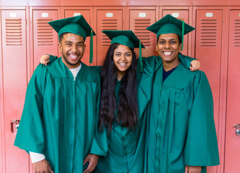 Three smiling students in green graduation gowns and caps standing in front of red lockers with arms around each other.