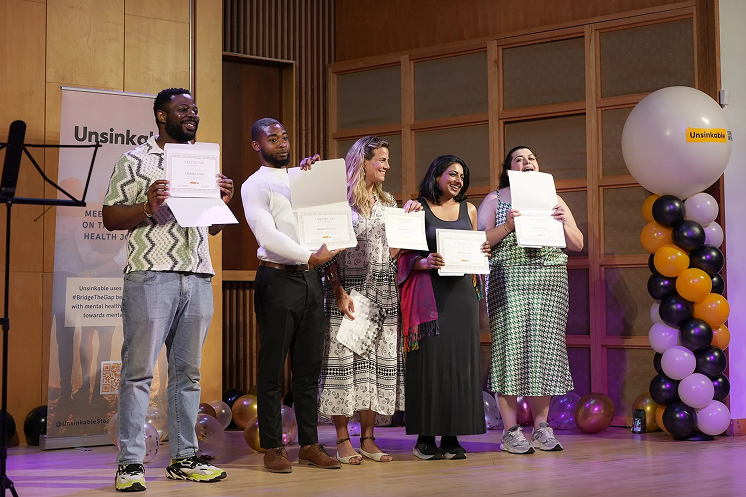 Five people standing on a stage holding certificates, smiling, with a backdrop of wooden panels and black, gold, and white balloons.