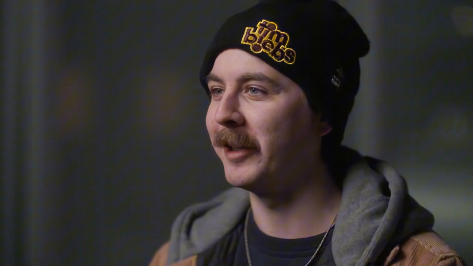 Person wearing a black beanie with yellow text and a gray hoodie under a brown jacket, looking slightly to the side.