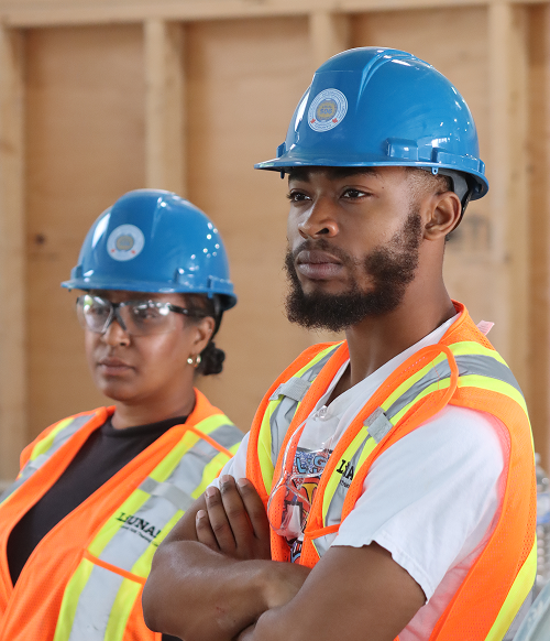 Two construction workers wearing blue helmets and orange safety vests indoors, one man with arms crossed and one woman with safety glasses.