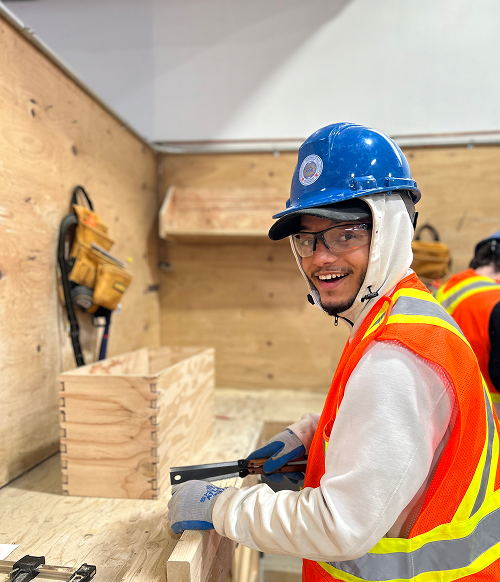 Smiling construction worker wearing a blue hard hat, safety glasses, white hoodie, and orange safety vest while working with wood.